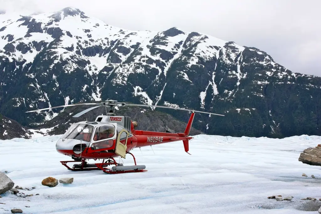 A red and white tour helicopter parked on the ice of Mendenhall Glacier below snowy peaks