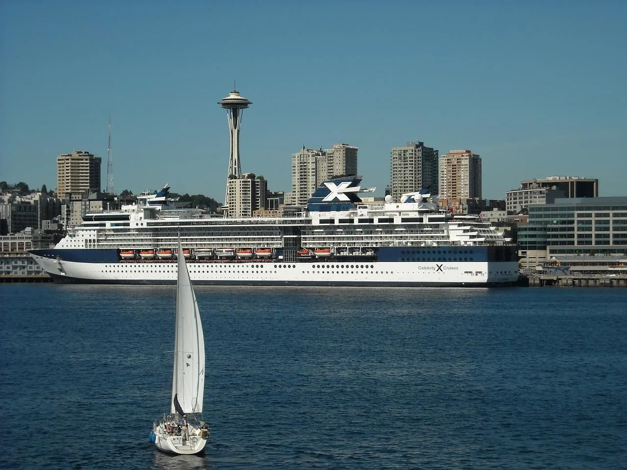 A white cruise ship sailing on Elliott Bay in front of the Space Needle, with a sailboat in the foreground, at the start of an Alaska cruise from Seattle
