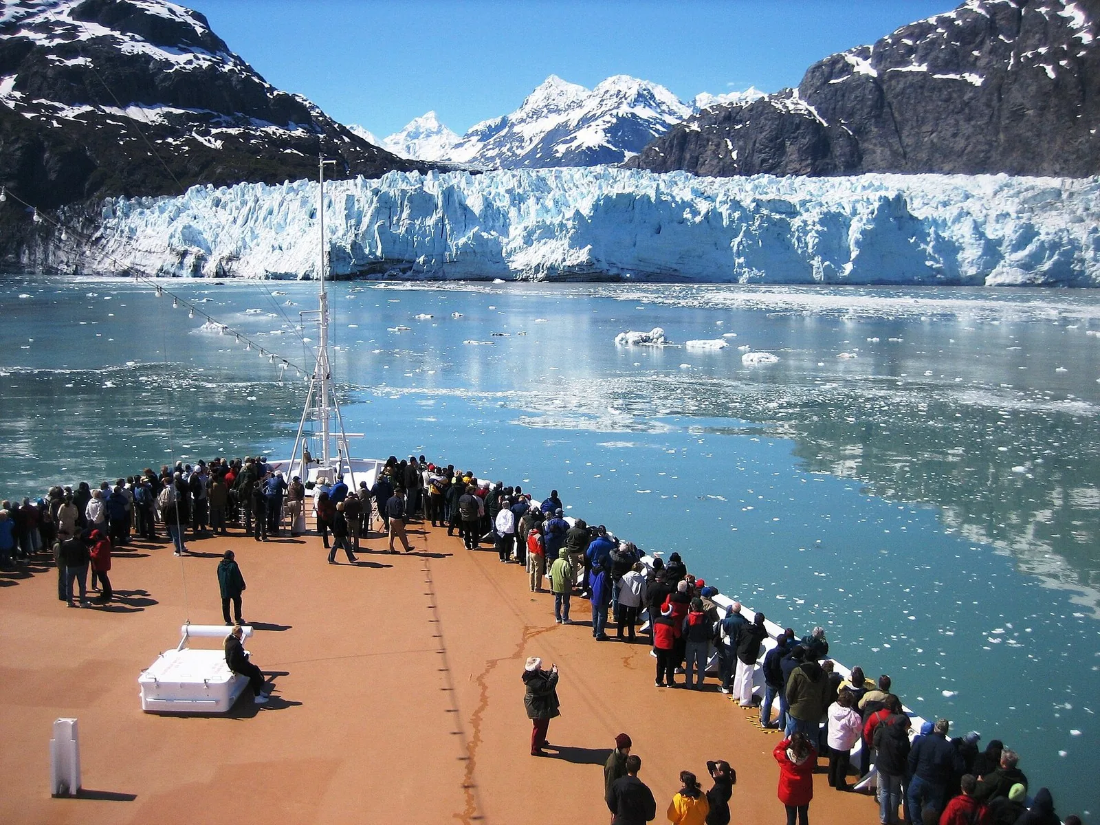 Cruise passengers in jackets and hats lining the bow as the ship faces a tidewater glacier in Glacier Bay National Park on an Alaska cruise
