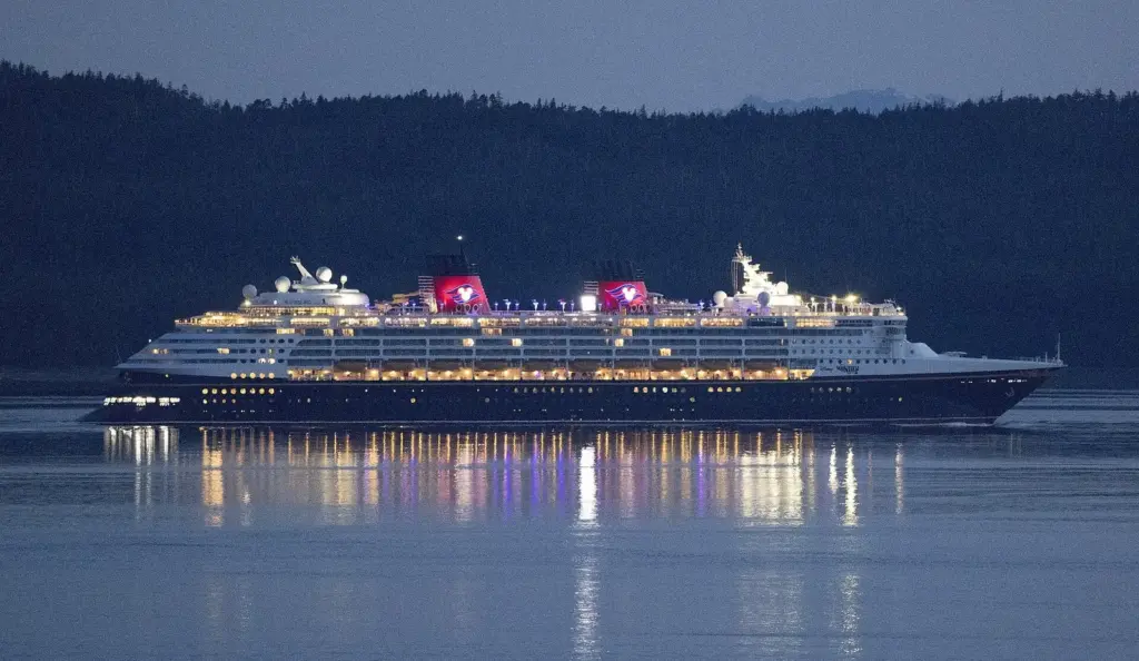 Disney Wonder lit up at dusk in Lynn Canal against dark forested mountains, its lights reflected in calm water