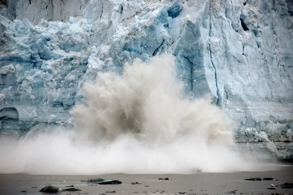 A huge spray of white water exploding from the base of a blue ice cliff as a section of the glacier face collapses