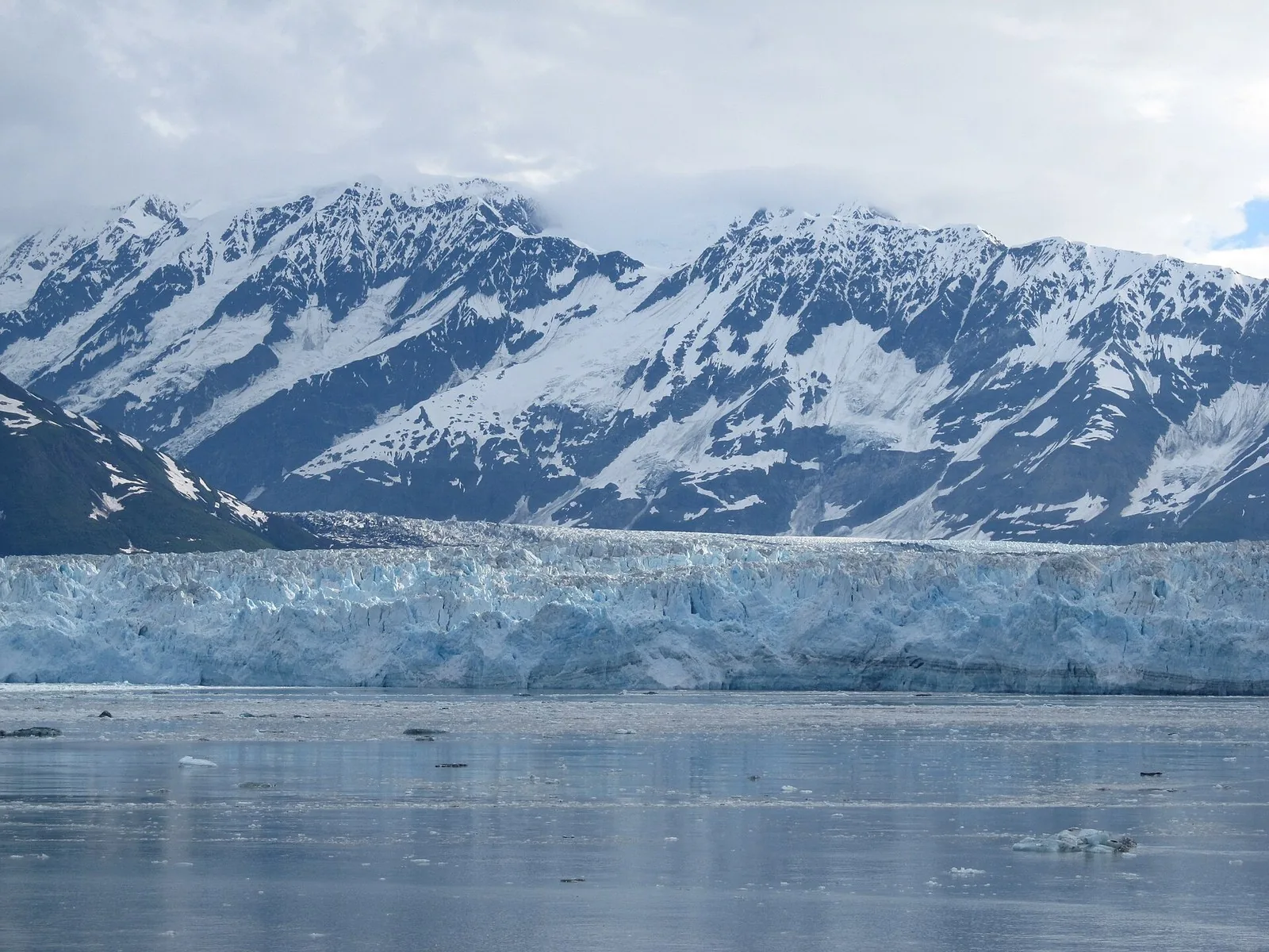 The long blue-white face of Hubbard Glacier across Disenchantment Bay, with snow-covered mountains rising behind and small icebergs on the water