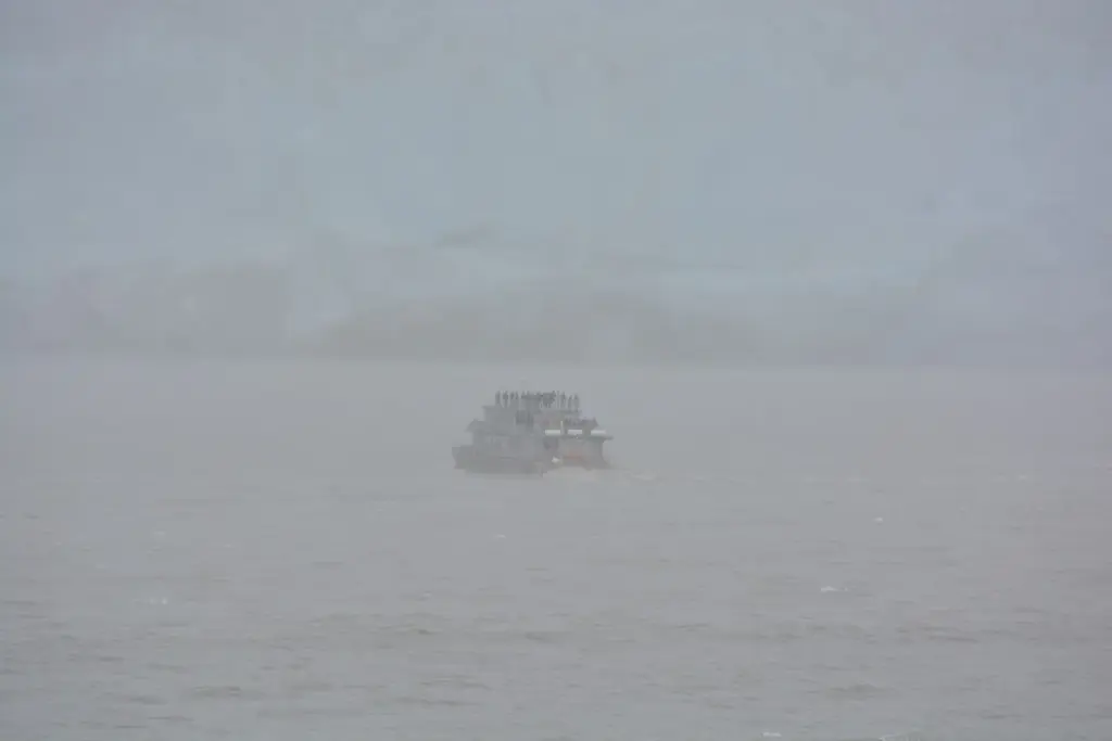 A small tour boat with passengers on its upper deck crossing gray water in thick fog near the ice