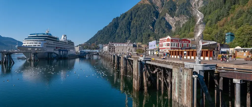 A cruise ship tied up along the long downtown Juneau cruise dock beneath forested Mount Roberts, with waterfront shops and a boardwalk