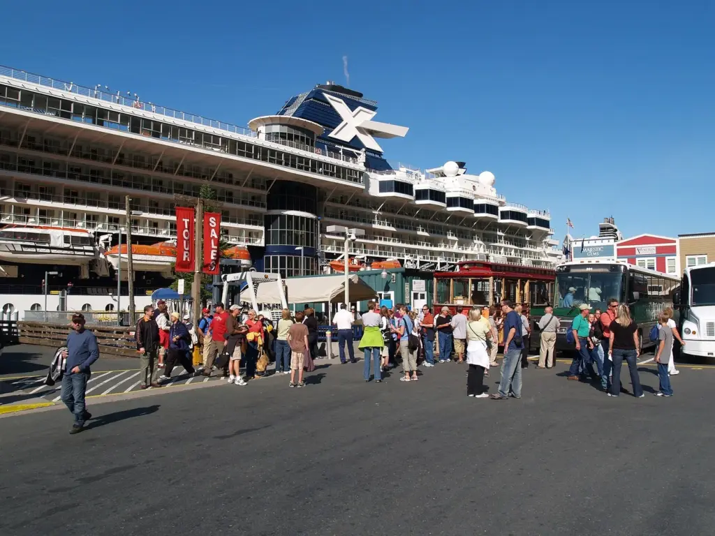 Cruise passengers in light jackets and caps gathering on the Juneau dock beside a ship and tour buses
