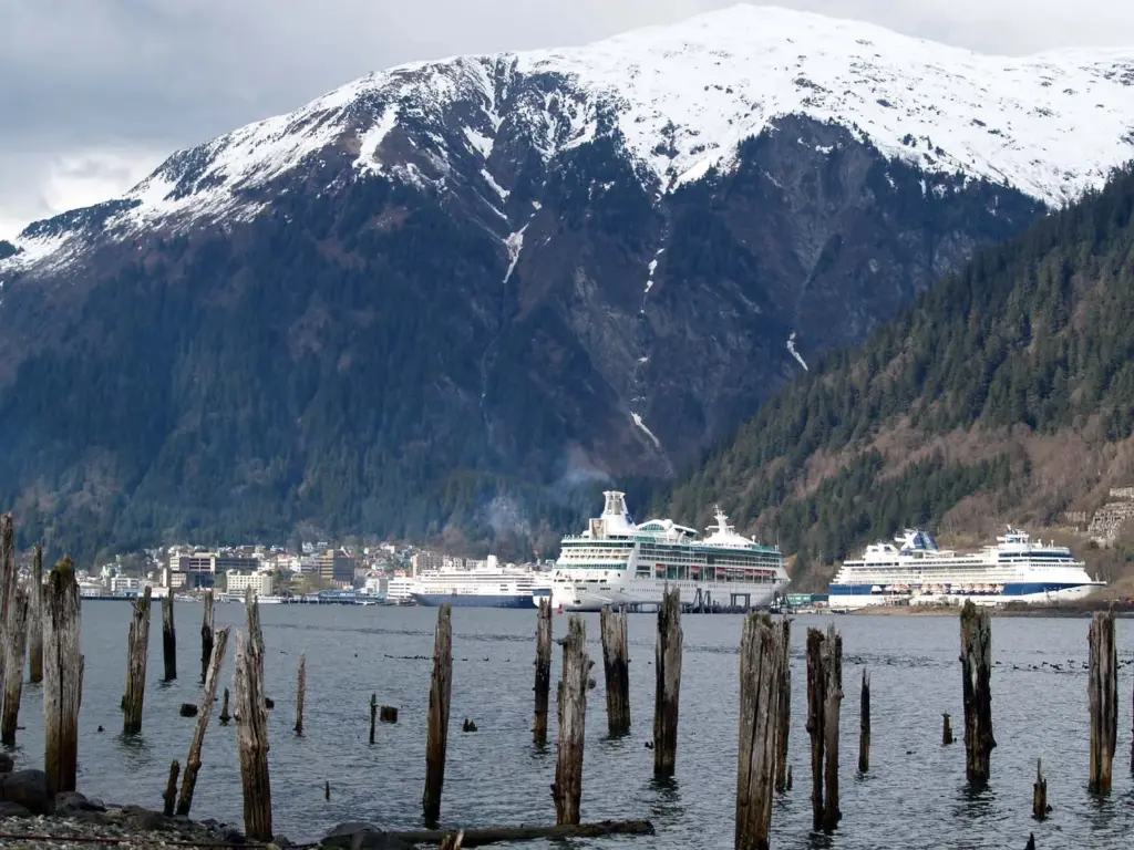 Three large cruise ships docked along the Juneau waterfront below snow-streaked Mount Juneau, seen past old wooden pilings on Douglas Island