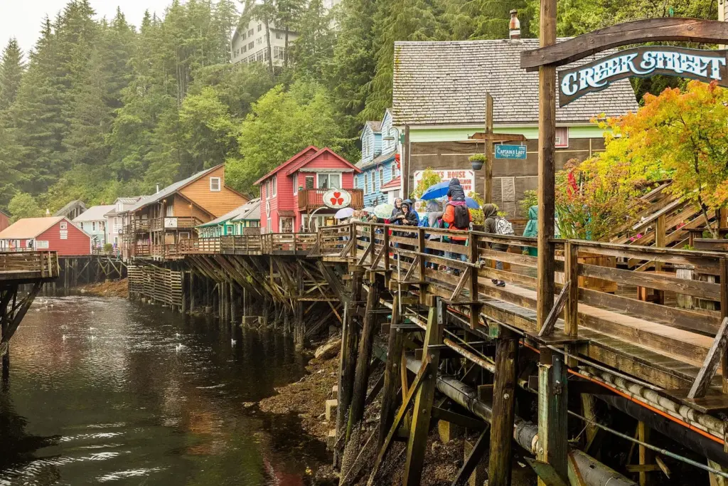 Cruise visitors with umbrellas and rain jackets on the boardwalk of Creek Street in Ketchikan, with colorful wooden buildings on pilings over the creek