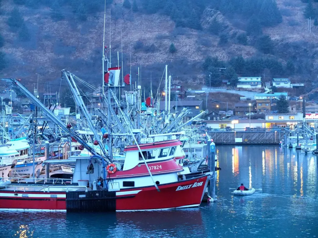 Commercial fishing boats crowded in the Kodiak harbor at dusk with the town lights behind