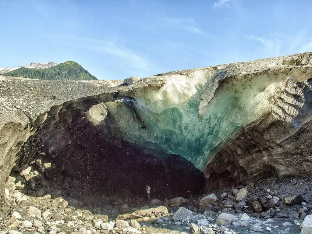 A wide blue ice cave opening beneath a thick, dirty layer of glacier ice and loose rock