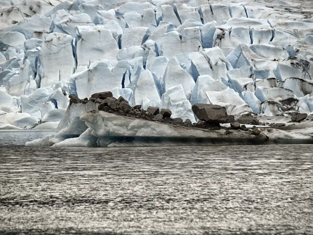 A dirty iceberg carrying dark moraine rocks, floating in front of a crevassed blue ice face