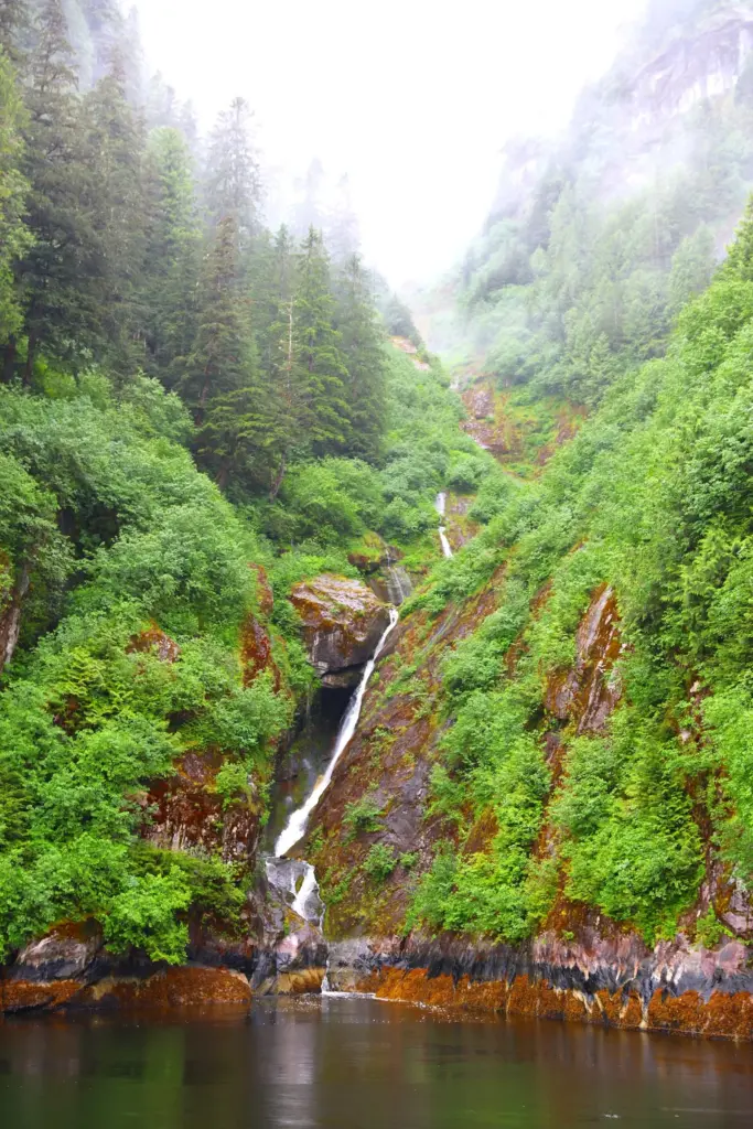 A waterfall pouring down a forested cleft between steep, mist-covered granite walls into the water in Misty Fjords National Monument