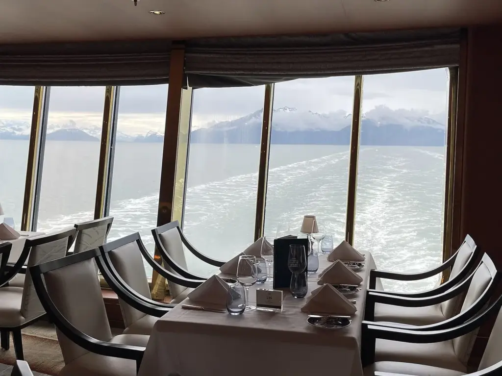 Tables set beside tall windows in the main dining room of Holland America's Noordam as the ship sails past mountains