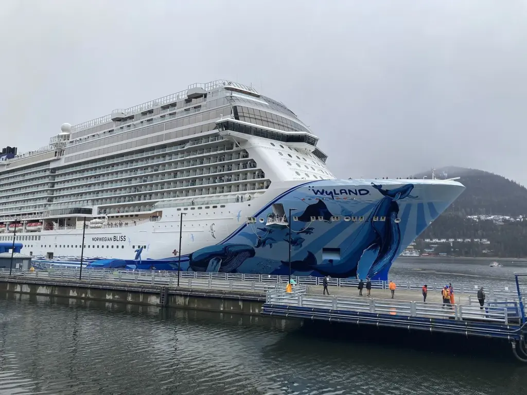 Norwegian Bliss, with an orca mural painted on its bow, docked at a Juneau cruise pier as passengers in rain jackets walk along the dock