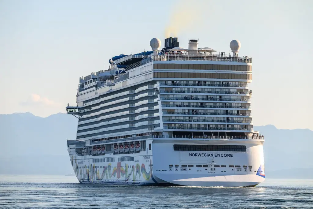 Norwegian Encore sailing away from Victoria's Ogden Point in the evening light, with low hills behind