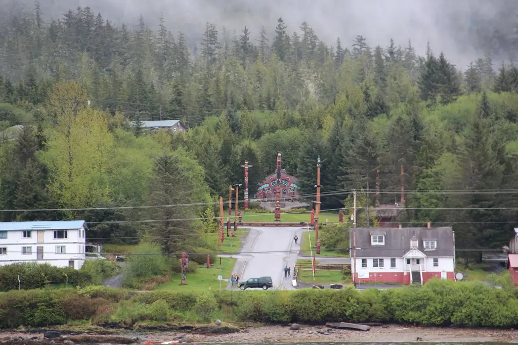 Totem poles and a painted clan house at the Saxman totem park, seen across the water below a forested hillside