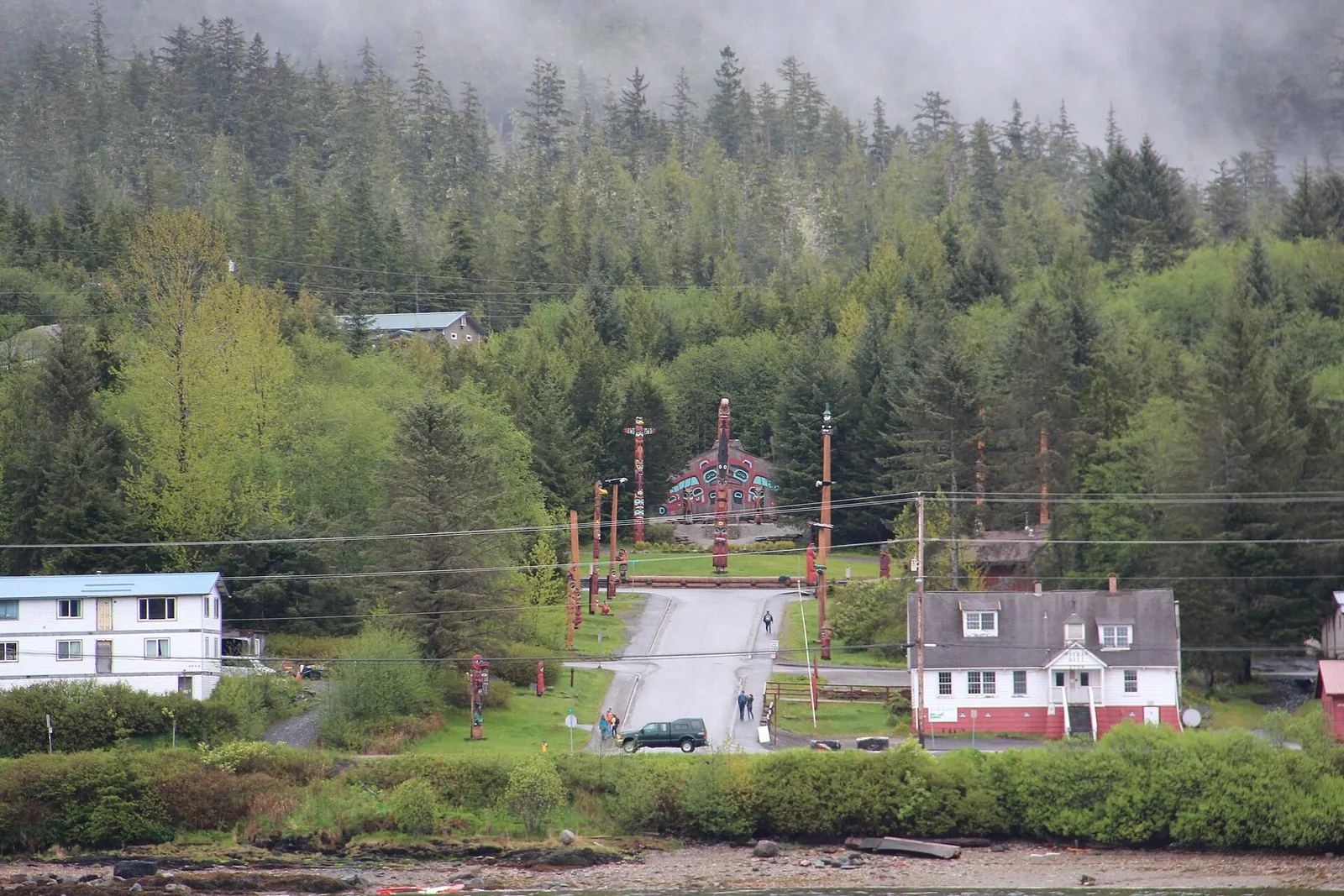 Totem poles and a painted clan house at the Saxman totem park near Ketchikan, seen across the water below a forested hill