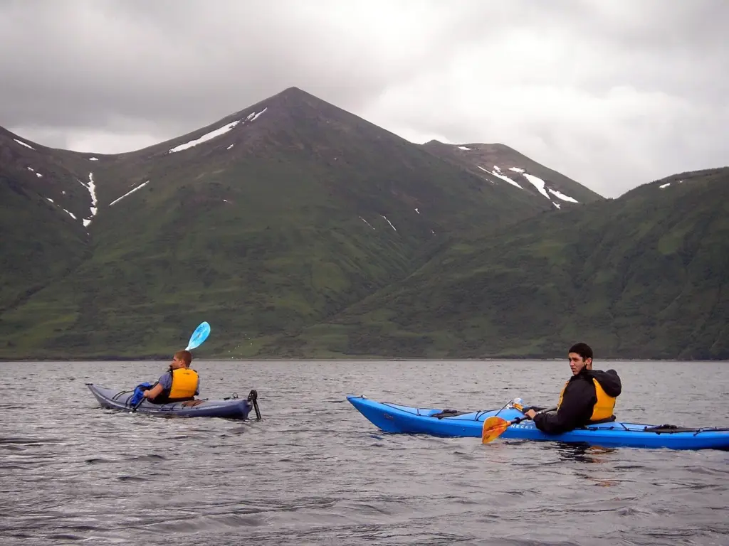 Two sea kayakers in life jackets paddling on gray water below green mountains in the Kodiak Archipelago