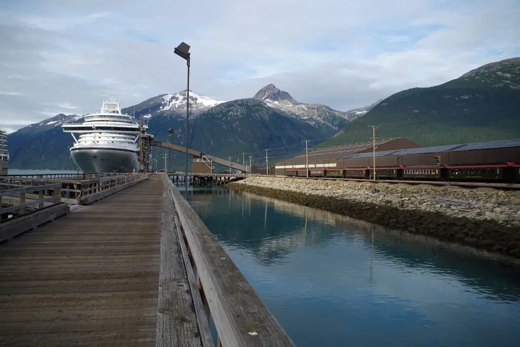 A large cruise ship at the end of a long Skagway dock, with White Pass railcars waiting alongside and snowy peaks behind