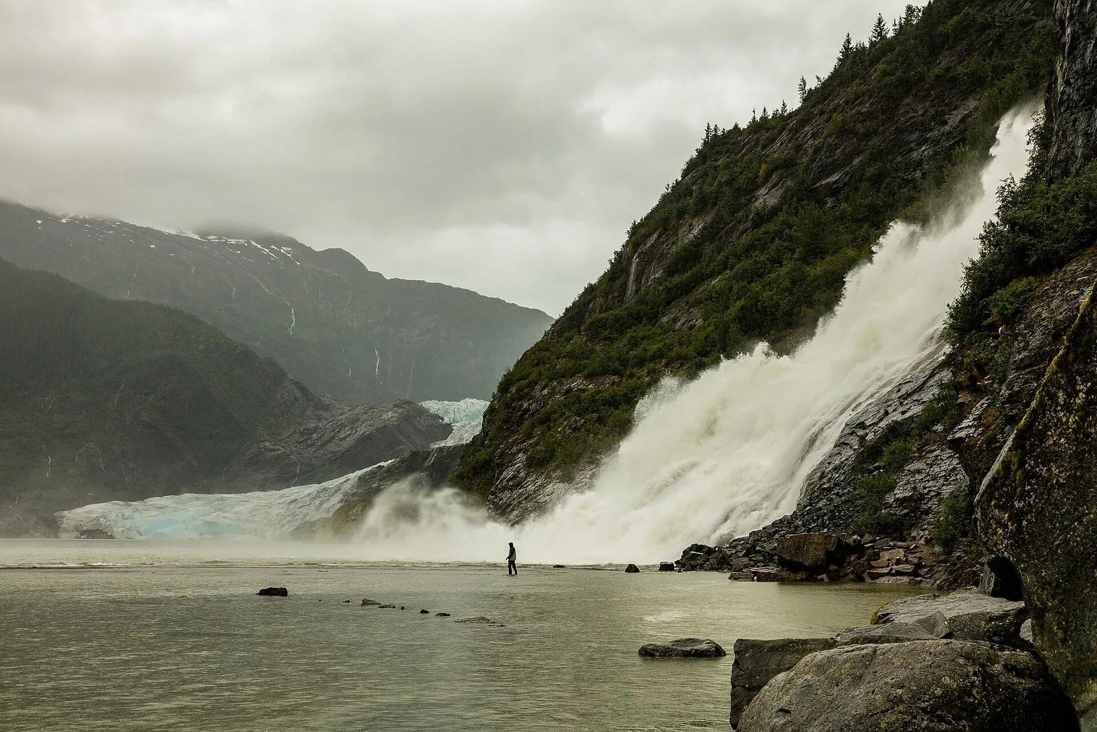 Nugget Falls pouring into Mendenhall Lake beside the blue ice of Mendenhall Glacier, with a lone visitor at the water's edge; the glacier tops most lists of things to do in Juneau
