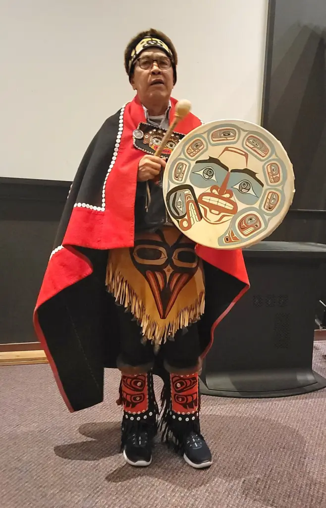 A Tlingit singer and drummer in a red and black button robe and carved headpiece, sharing Alaska Native culture at the top of the Goldbelt Tram in Juneau