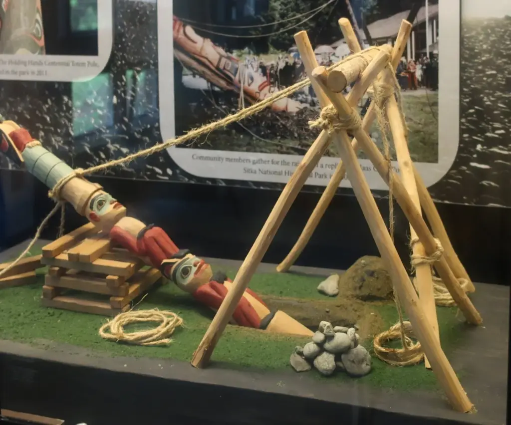A museum model showing a totem pole being raised with ropes and a wooden A-frame