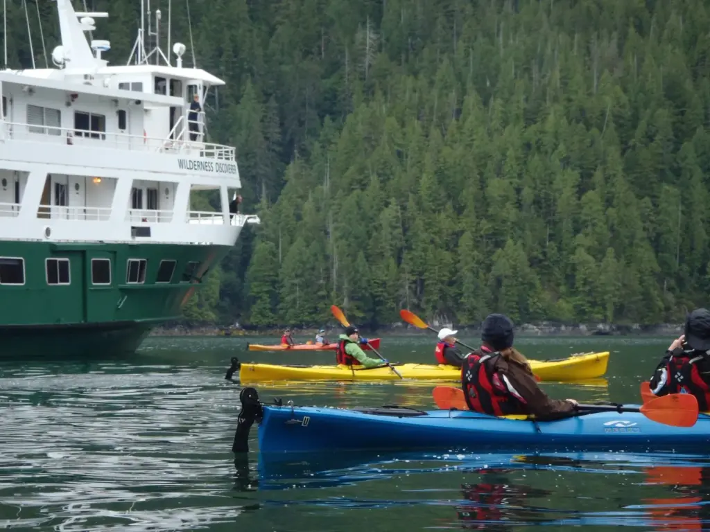 Kayakers in yellow and blue boats paddling beside the green-hulled Wilderness Discoverer below forested slopes