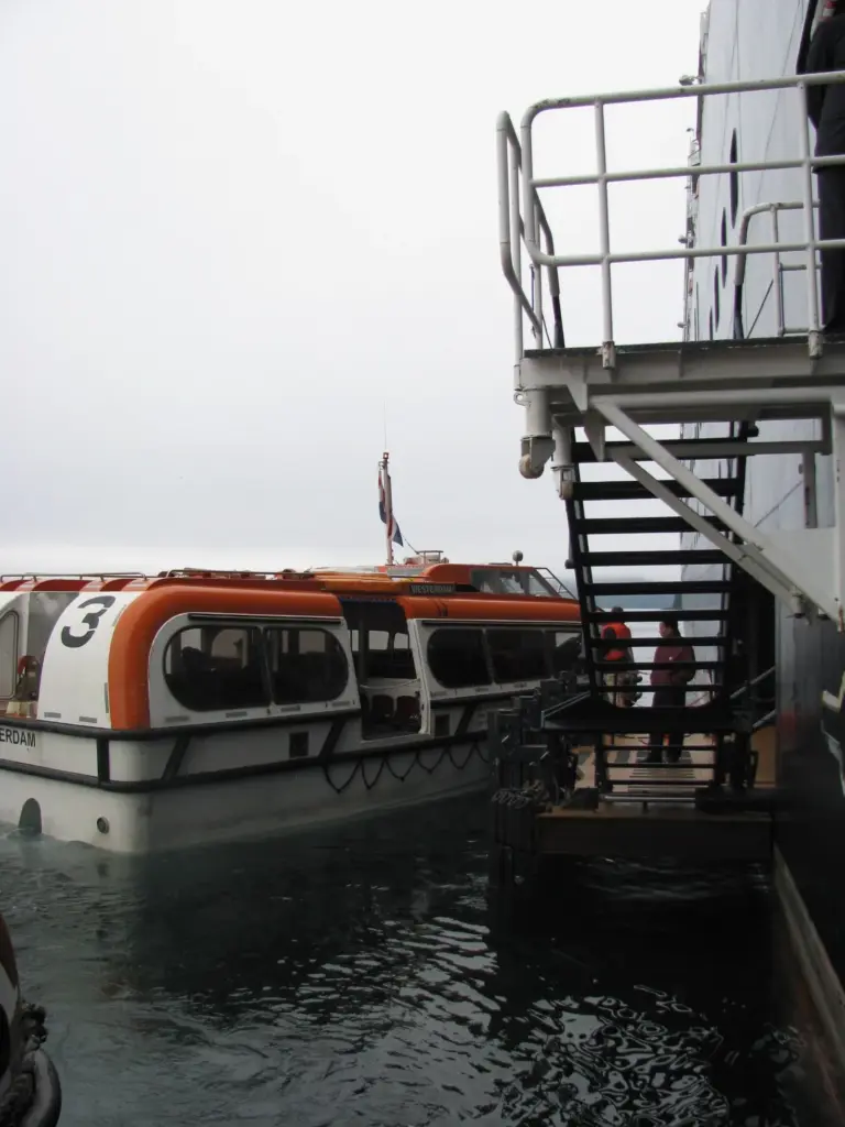 An orange and white ship's tender boat tied up beside a boarding platform and staircase on the side of a cruise ship