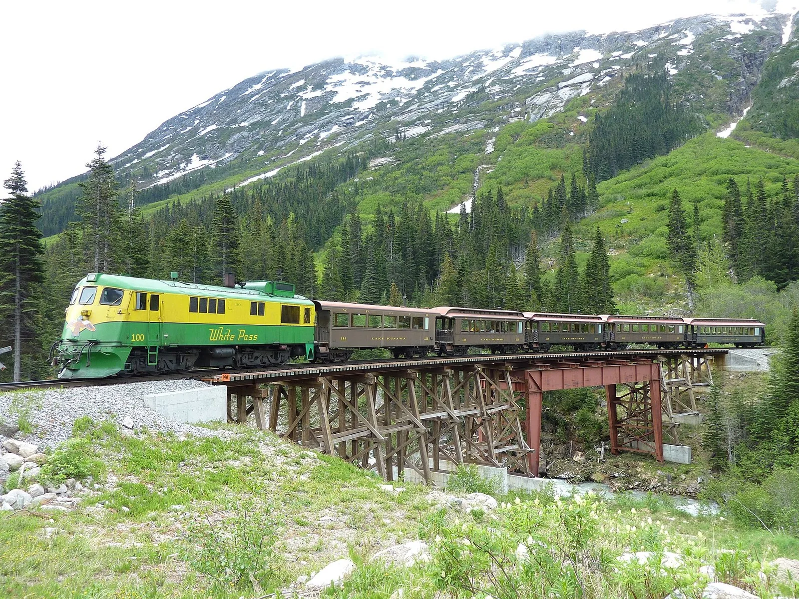 A green and yellow White Pass Railroad diesel pulling vintage passenger cars across a bridge below snow-streaked mountains