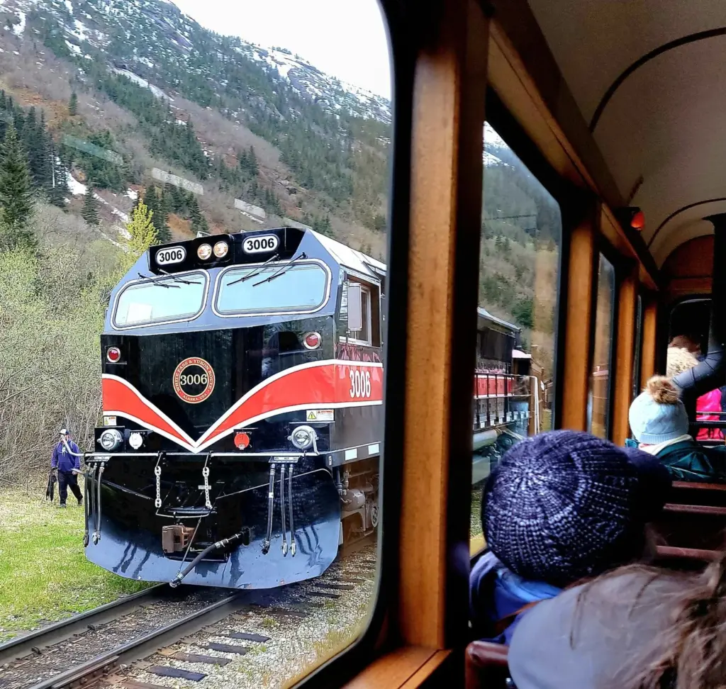 A passenger in a knit hat watching a red, white and blue diesel locomotive pass the window of a vintage wooden rail car