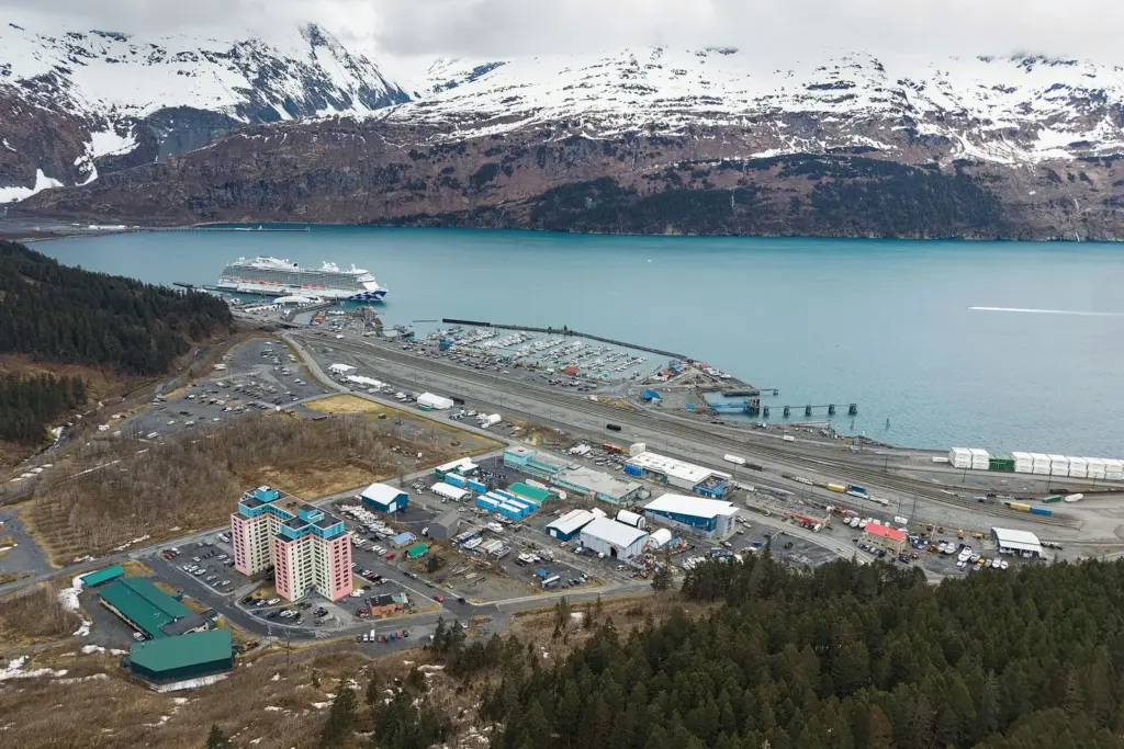 Aerial view of Whittier, Alaska, with a large cruise ship at the dock, the rail yard and the Begich Towers building below snowy mountains