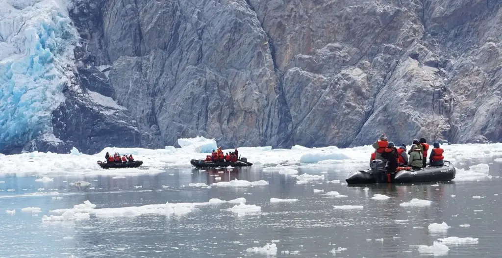 Three inflatable Zodiacs carrying passengers in orange jackets through floating ice toward a blue glacier face